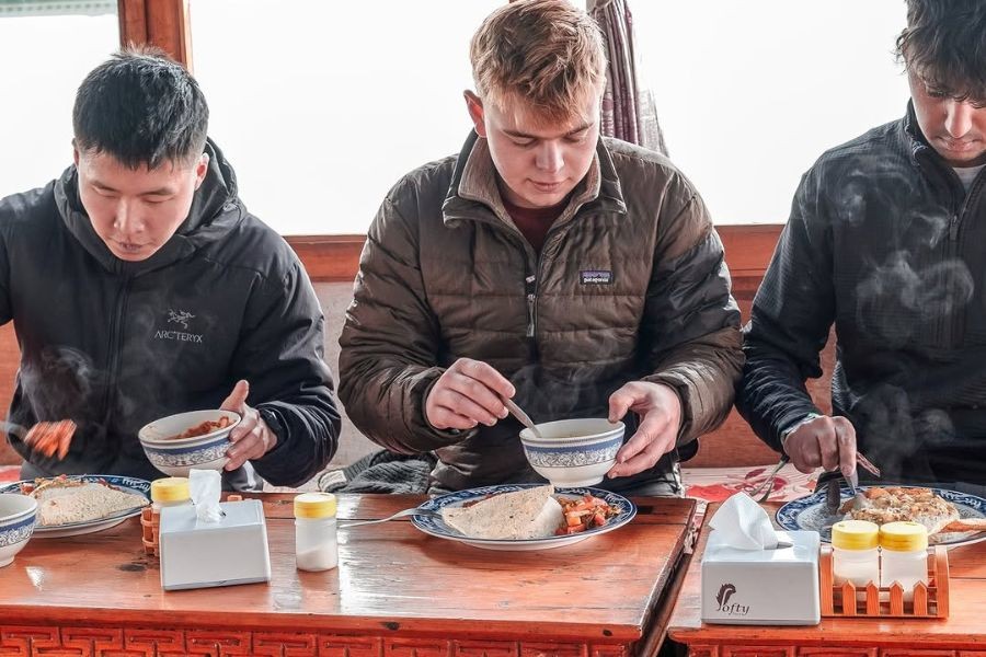 Three trekkers eating breakfast at wooden table inside mountain lodge